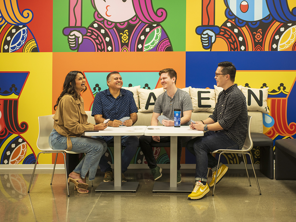 MobilityWare team members sit at a table playing cards and laughing in front of vibrant graphic wall and lettered pillows that read “game on.”